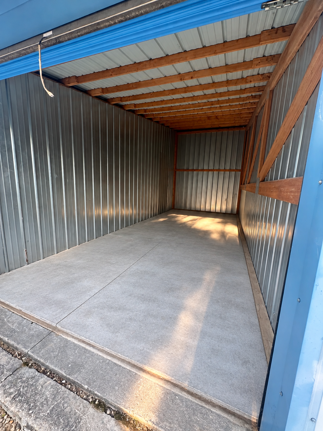 Interior of an open storage unit with a blue roll-up door, galvanized metal walls, wood-framed roof, and a smooth concrete floor.